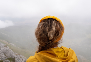 Photo from the back of a Caucasian blonde female hiker, dressed in yellow looking at the landscape in a mountainous area with fog