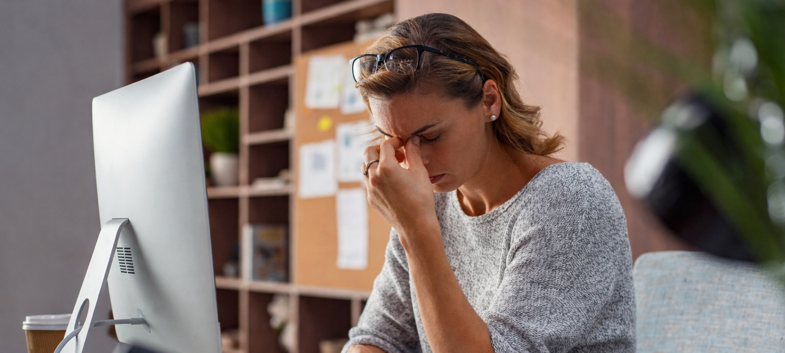 Eine Frau sitzt erschöpft an ihrem Arbeitsplatz, die Augen geschlossen, eine Hand an der Stirn, vor ihr Computer und Unterlagen. Das Bild vermittelt die Botschaft von Erschöpfung und Reslienz: Arbeitsfähigkeit gerät nicht durch mangelnden Einsatz in Gefahr, sondern durch dauerhaft hohe Anforderungen und fehlende Regulation. Resilienz zeigt sich hier nicht im Durchhalten, sondern im ernsthaften Wahrnehmen von Erschöpfung als Warnsignal und im Bedarf nach bewusster Selbststeuerung, Abgrenzung und nachhaltiger Anpassung im Arbeitskontext.