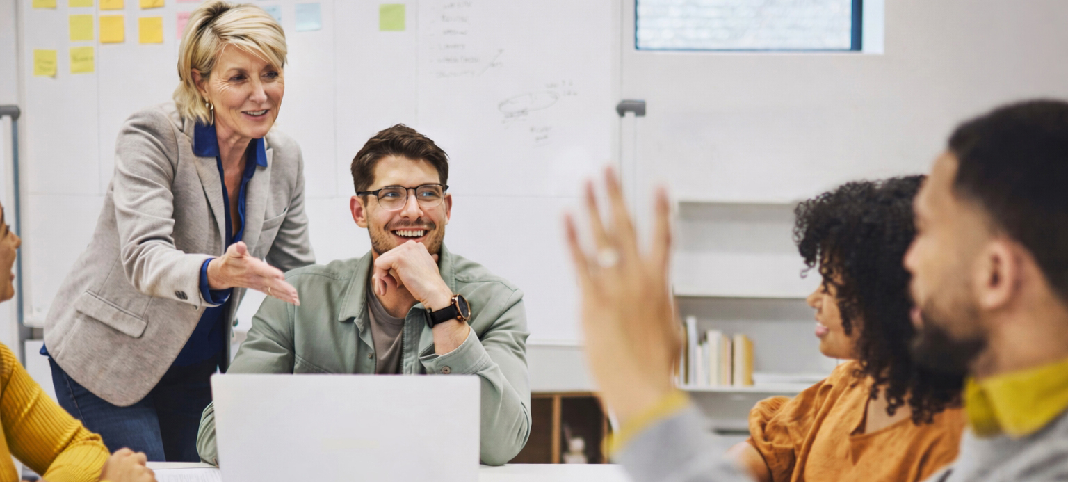 Eine Führungskraft steht in einem Teammeeting und bindet die Mitarbeitenden aktiv in den Austausch ein, während mehrere Teammitglieder aufmerksam zuhören und sich beteiligen. Das Bild vermittelt die Botschaft von Führungskultur und organisationale Resilienz: Eine offene, dialogorientierte Führungskultur schafft psychologische Sicherheit, fördert Lernen und Beteiligung und ermöglicht es Organisationen, mit Unsicherheit konstruktiv umzugehen. Es verdeutlicht, dass organisationale Resilienz weniger durch formale Leitbilder entsteht als durch gelebte Führung im Arbeitsalltag.