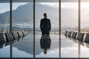 Ein Mann steht allein in einem modernen Konferenzraum vor einer großen Fensterfront und blickt auf die Stadt und die Berge. Das Bild vermittelt die Botschaft der Normative Dimension von Resilienz: Resilienz im Führungskontext ist nicht wertneutral, sondern mit Erwartungen, Leistungsnormen und impliziten Vorstellungen von „angemessener“ Anpassung verbunden. Es symbolisiert die Verantwortung von Führung, kritisch zu reflektieren, woran Anpassung eingefordert wird, für wen sie gilt und wann Resilienz auch bedeutet, Grenzen zu setzen oder bestehende Bedingungen infrage zu stellen.